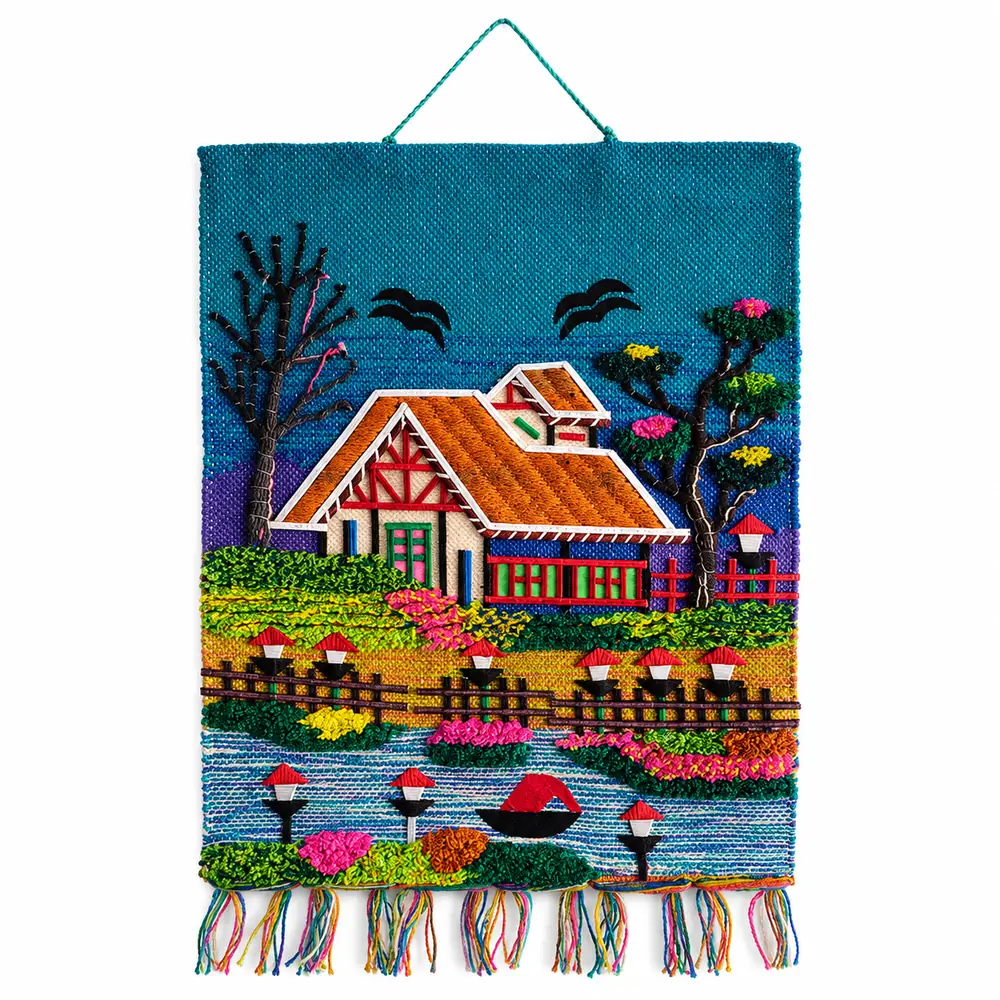 Handmade woven wall hanging with a raised yarn cottage, lakeside lamp posts, boat and colourful tassel fringe on a turquoise background