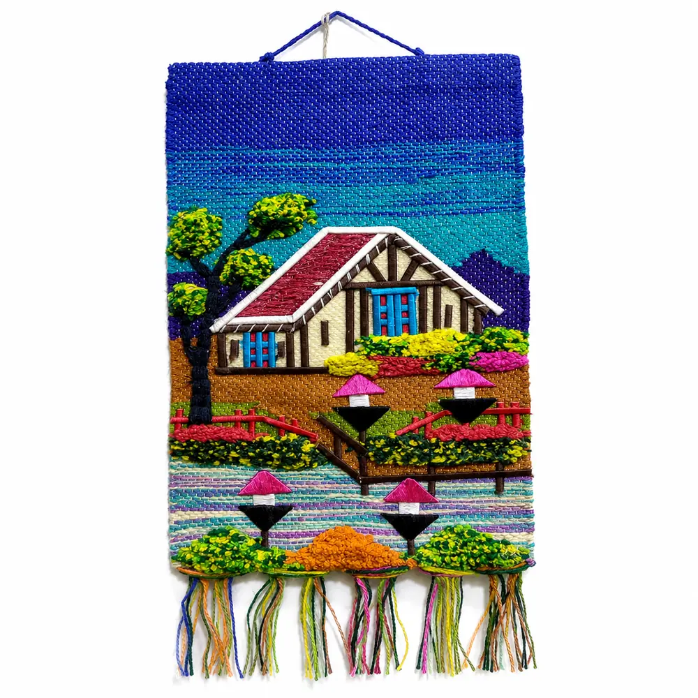 Handwoven wall hanging with a blue sky, a single embroidered cottage with red roof, a tree, fence and colourful tufted bushes with tassel fringe