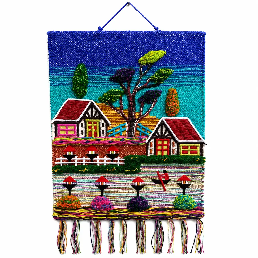 Woven village scene wall hanging in blue and turquoise with two cottages, a large tree, a white fence, a rowing boat and pom-pom bushes