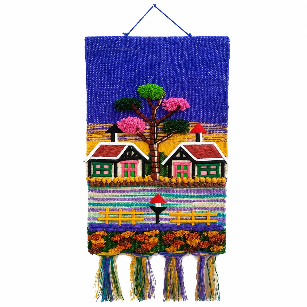 Blue and yellow woven wall hanging with two cottages, a pink-blossom tree, yellow fences and orange tufted flowers, finished with thick tassels