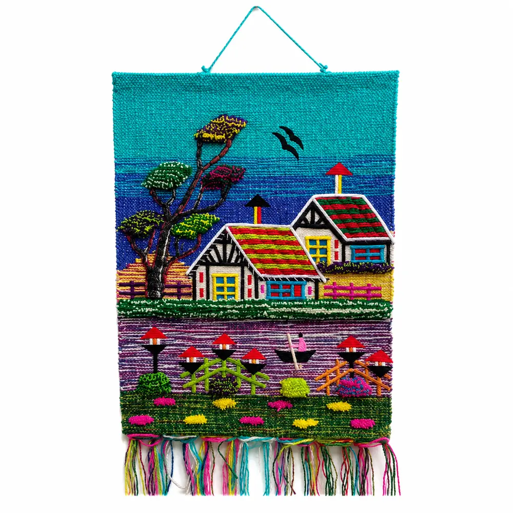 Turquoise and blue woven wall hanging with two cottages with striped roofs, a tree, birds, a boat on purple water and a green field with tassels